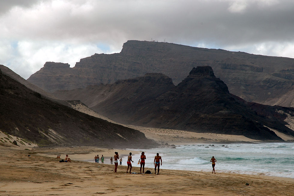 Praia deserta com banhistas, mar agitado e montanhas escuras sob céu nublado e dramático.