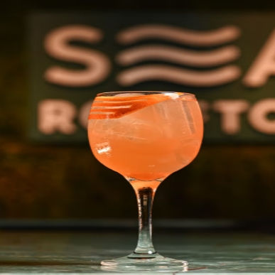 Taça de coquetel laranja com gelo sobre o balcão. Ao fundo, placa desfocada com letras brancas em fundo verde, sugerindo restaurante.
