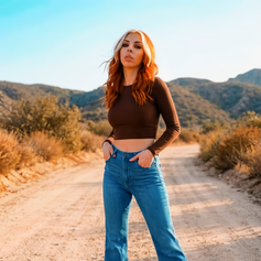 Ari with copper hair, poses in brown ribbed crop top and high waisted blue flare jeans on a dirt road.