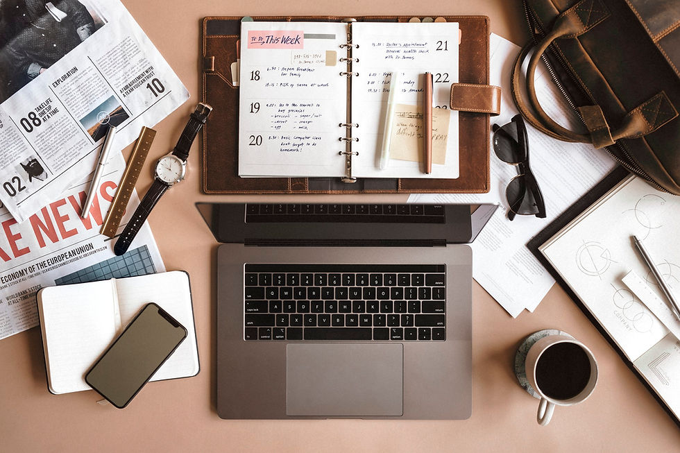 Top view of a workspace with an open planner, laptop, smartphone, watch, and coffee. Papers, sunglasses, and leather bag in the background.