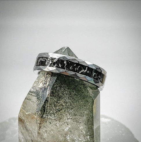 A hammered tungsten ring with an inlay of genuine meteorite fragments, displayed on top of a clear quartz crystal. The dark inlay contrasts against the faceted metal edges, highlighting the cosmic-inspired design.