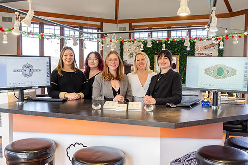 A team of five smiling jewelry designers and consultants stand behind a sleek design counter inside a jewelry store. The workspace features two computer monitors displaying intricate custom ring designs. The store is well-lit, with modern decor, festive garland, and jewelry showcases in the background. Two jewelry trays with rings are displayed on the counter. The team members are dressed in stylish, professional attire, exuding a welcoming and creative atmosphere.