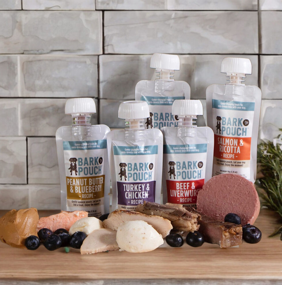 A variety pack of Bark Pouch liquid dog treats in front of ingredients like peanut butter, salmon, chicken, and liverwurst on a cutting board.