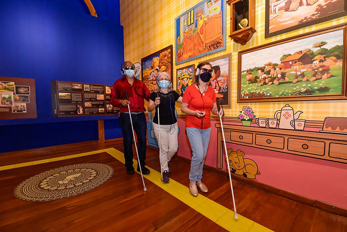 Na Pinacoteca Municipal de Jundiaí, em São Paulo, três pessoas com deficiência visual caminham juntas pelo espaço expositivo, utilizando bengalas brancas. Elas usam máscaras de proteção e óculos escuros. O piso é de madeira e conta com faixa de piso tátil em relevo, na cor amarela.