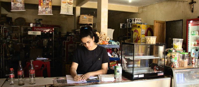 Mulher branca, com cabelos pretos amarrados e vestindo blusa preta, se apoia em um balcão de bege, com os braços sobre uma folha de papel na qual escreve à caneta com a mão direita. Ela está em um mercado, com expositores, geladeiras, banners e produtos para venda.