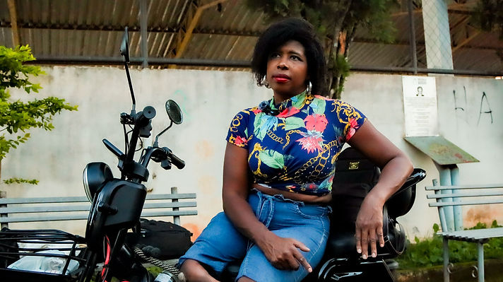 Mulher negra adulta sentada em triciclo preto ao ar livre. Usa blusa azul estampada e bermuda jeans, com brincos grandes. Olha para cima com expressão atenta. Ao fundo, banco, muro claro, cobertura metálica e árvores, em cena diurna.