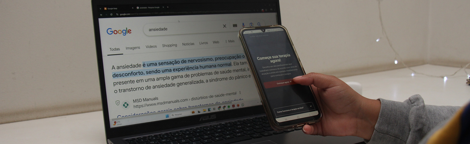 Para Todos Verem: Mão negra, que está apoiada em uma mesa branca, segurando um celular. Na tela é possível ler: “Comece sua terapia agora”. Atrás da mão, há um notebook em cima da mesa com a tela aberta no Google, exibindo resultado de pesquisa sobre a palavra “ansiedade”.