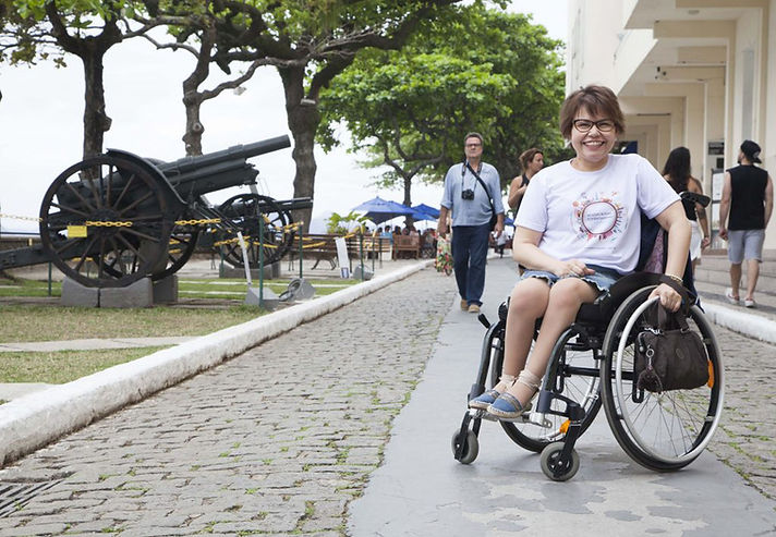Área externa do Forte de Copacabana, no Rio de Janeiro. Em primeiro plano, uma mulher sentada sobre uma cadeira de rodas manual está parada sobre uma faixa de circulação lisa inserida em meio ao calçamento de pedras.