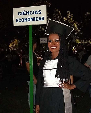 Cinara, mulher preta com tranças pretas com detalhes brancos, usando beca de formatura preta e branca parada em frente a uma placa indicando o curso de Ciências Econômicas. Na foto ela aparece sorridente.