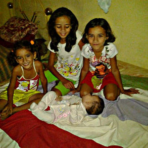 Quatro crianças estão em cima de uma cama, as três mais velhas estão sentadas e no meio delas está uma neném, deitada. Todas sorriem.