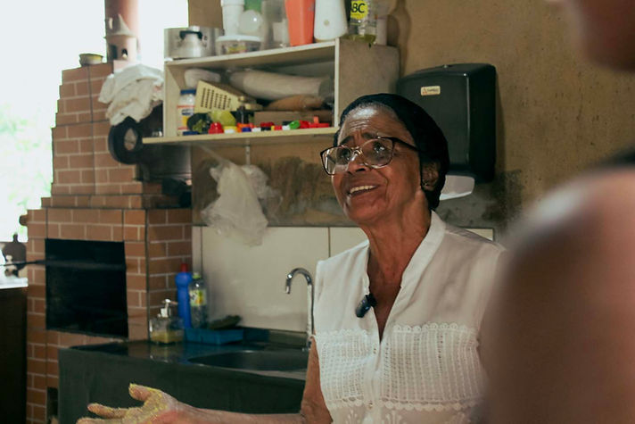 Clemilda sorri, sua mão está amarela por conta dos grãos de fubá de milho. Ela está de óculos, touca de cozinha e blusa branca.