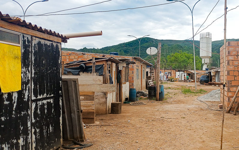 Imagem do Terreno da ocupação Chico Rei em que podemos ver 7 casas simples, de madeira, e tijolos, sem reboco, alinhadas à esquerda, à direita e ao fundo da imagem, e ao centro uma grande parte de chão de terra batida ocupa a imagem.
