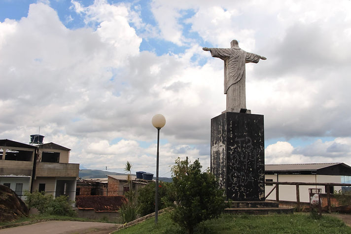 Estátua de Cristo de braços abertos, vista de costas, no alto do bairro Cabanas. A escultura está inserida em uma praça, com casas do bairro ao redor e o céu nublado ao fundo.