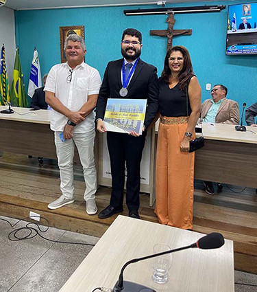 No centro da imagem, Dener, um homem branco de cabelo curto, barba e óculos, está de terno preto e com uma medalha em volta do pescoço enquanto carrega uma placa. Ao lado dele, há duas pessoas: do lado direito da imagem, está uma mulher usando uma blusa preta e uma calça laranja, à esquerda, está um homem todo vestido de branco. Todos estão sorridentes.