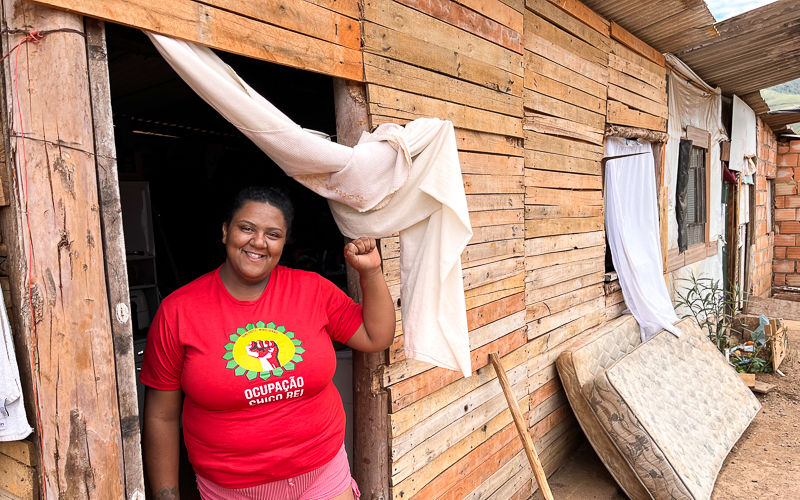 Uma mulher negra de cabelos pretos está na porta de sua casa. Ela usa uma camiseta vermelha com o símbolo de luta do movimento da ocupação Chico Rei e um short rosa. Ela sorri para a foto e faz o gesto de luta com as mãos, com o punho fechado e erguido.