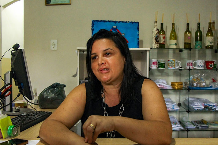 Mulher branca adulta de cabelos pretos lisos sentada atrás de mesa com computador e objetos de escritório. Ao fundo, prateleiras com itens decorativos e quadros na parede. Ambiente interno organizado.