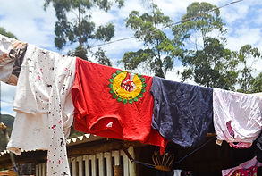 Cinco peças de roupas estão penduradas em um varal na Ocupação Chico Rei, uma delas é uma camisa vermelha com o símbolo do coletivo: uma mão de punhos fechados que representa luta e resistência. Ao fundo, é possível avistar um pouco do céu - azul e com algumas nuvens - e da copa de uma árvore.