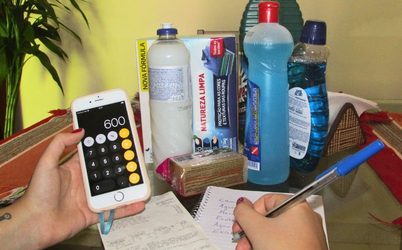 Mãos de uma mulher branca com unhas vermelhas, segurando uma caneta e um telefone celular aberto em um aplicativo de calculadora sobre uma nota fiscal e um papel com anotações que estão em cima de uma mesa. Ao fundo, na mesma mesa, há produtos de limpeza.