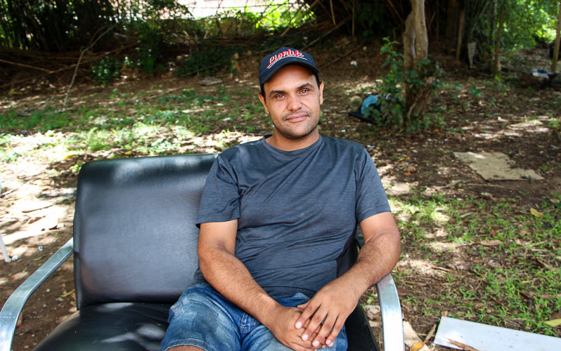 Um homem pardo, que usa boné azul, uma camisa cinza e um short jeans, está em meio a um gramado, sentado em uma cadeira preta e encosta seu braço esquerdo no apoio do assento.
