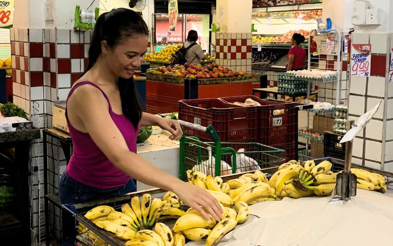 Josiane Machado Mulher de cabelos pretos amarrados, vestindo camiseta de cor roxa e calças jeans. Ela está em um sacolão com as mãos prestes a pegar um cacho de banana. Ao fundo pessoas, caixotes, frutas, placas de preços e um carrinho de compras.