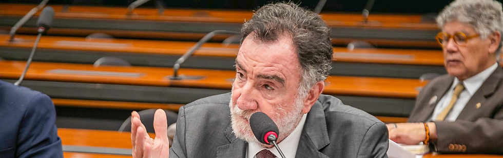 A foto mostra um homem de pele branca, de cabelos grisalhos, barba branca e terno cinza em frente ao microfone com a mão levantada. Ao fundo há um outro homem com as mesmas descrições, com a mão em cima da mesa e a cor da pele parda.