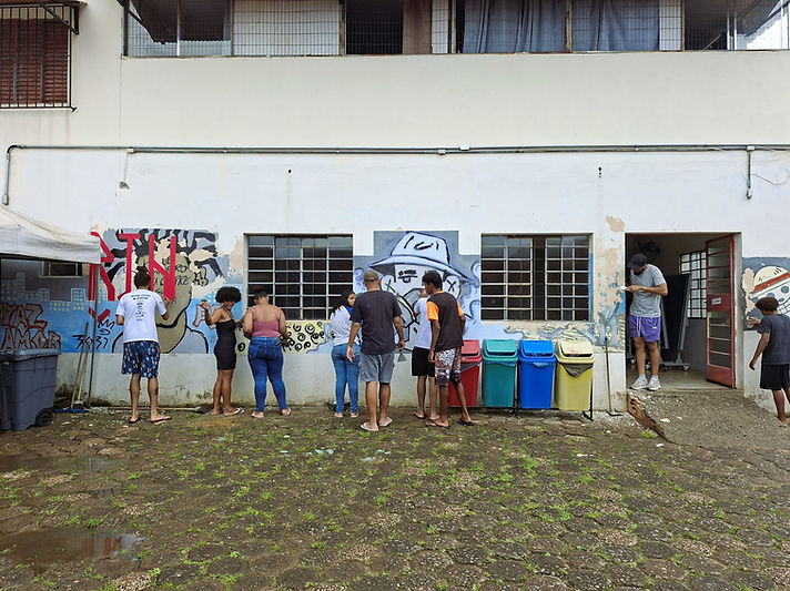 Jovens e adultos pintam murais coloridos em uma parede branca, ao lado de quatro lixeiras de coleta seletiva.