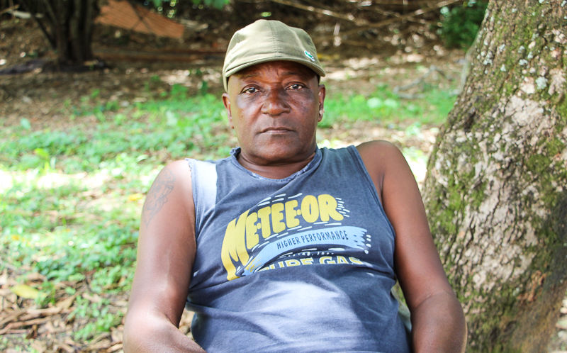 Um homem negro usa um boné verde e uma camiseta azul com inscritos em amarelo: “Metee-or”. Ele está em uma área gramada, na qual se avista parte do tronco de uma árvore.