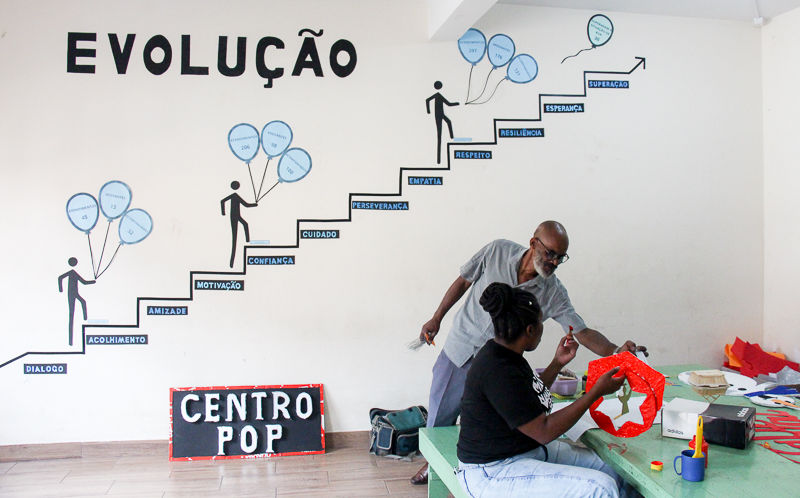 No primeiro plano, à direita, um homem negro careca, que usa óculos de grau, veste uma camisa cinza e uma calça azul, ajuda uma mulher negra em sua pintura. A mulher, de cabelos pretos, presos em um coque, está sentada em um banco verde, diante de uma mesa também verde, e pinta uma peça com tinta vermelha. Ao fundo, em uma parede branca, há uma ilustração de pessoas subindo degraus com balões na mão. Em cada degrau há inscrita uma palavra de apoio, como "motivação"e "confiança" . No último degrau, uma seta aponta para cima. À esquerda, a palavra “evolução” está gravada em grandes proporções. Encostada na mesma parede, está uma placa preta com bordas vermelhas com “Centro POP” escrito em branco.