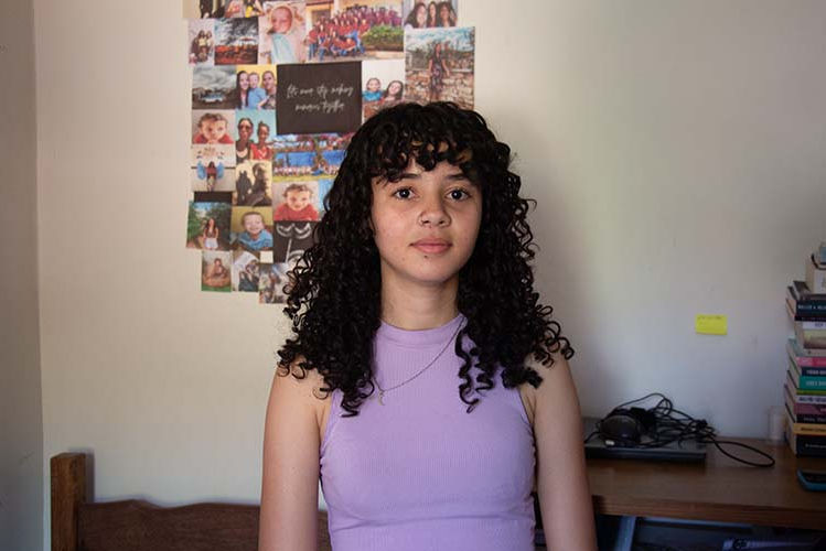 Ana Cláudia, jovem de cabelo cacheado preto e blusa lilás, está com o rosto pouco inclinado para a esquerda parada em frente a um mural de fotos. Ao fundo há também uma mesa com notebook, carregador e livros.