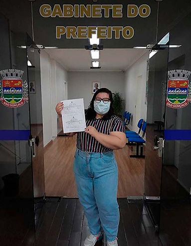 Bruna, uma mulher branca de cabelo liso castanho, está usando uma blusa listrada, uma calça jeans clara e máscara segurando um documento em frente ao Gabinete do Prefeito. Nas portas é possível ver o símbolo da Prefeitura de São Gonçalo.