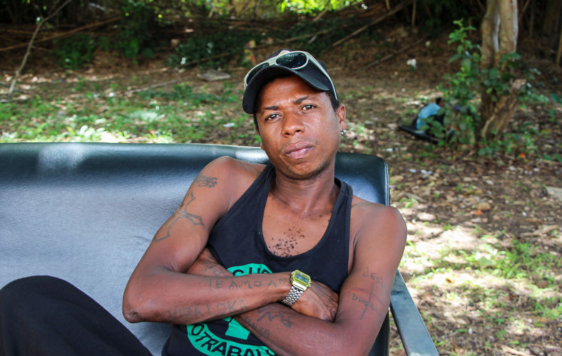 Um homem usa uma camiseta preta, um boné preto com um óculos cinza acima e um relógio no pulso esquerdo. Ele está sentado em uma cadeira, no meio de um gramado, com os braços cruzados e encara a câmera.
