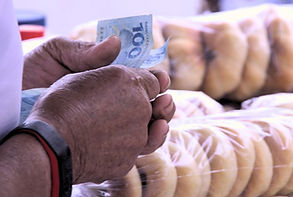 Mãos e busto de uma pessoa vestindo uma blusa branca. Ela segura duas notas de R$ 100. Ao fundo, pacotes de rosquinhas