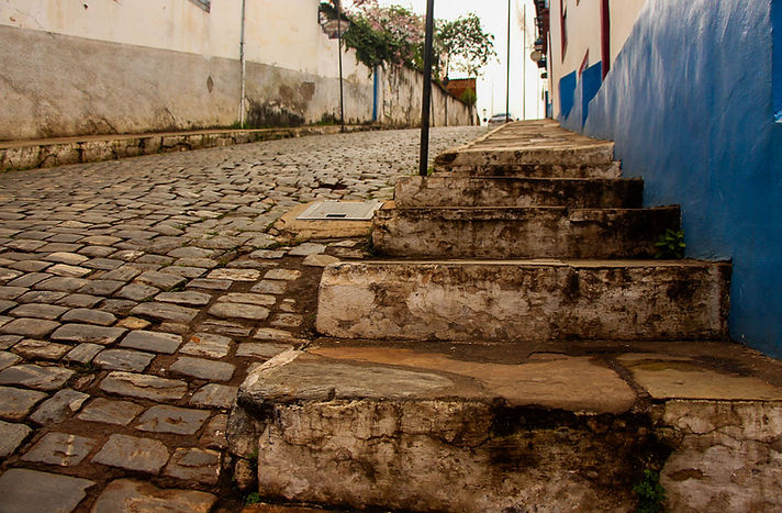Rua Dom Silvério em subida, com calçamento de pedras irregulares. À direita, degraus de pedra sem corrimão junto a parede azul e branca. Poste metálico no centro e via desnivelada à esquerda. A imagem evidencia barreiras de mobilidade no Centro Histórico.