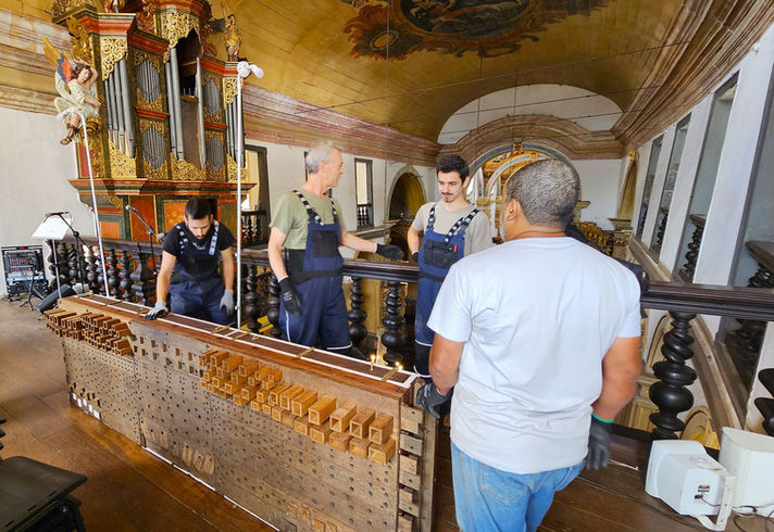 Quatro homens trabalham juntos em um grande instrumento musical de madeira. Eles vestem roupas de trabalho, como macacões, camisetas e luvas.