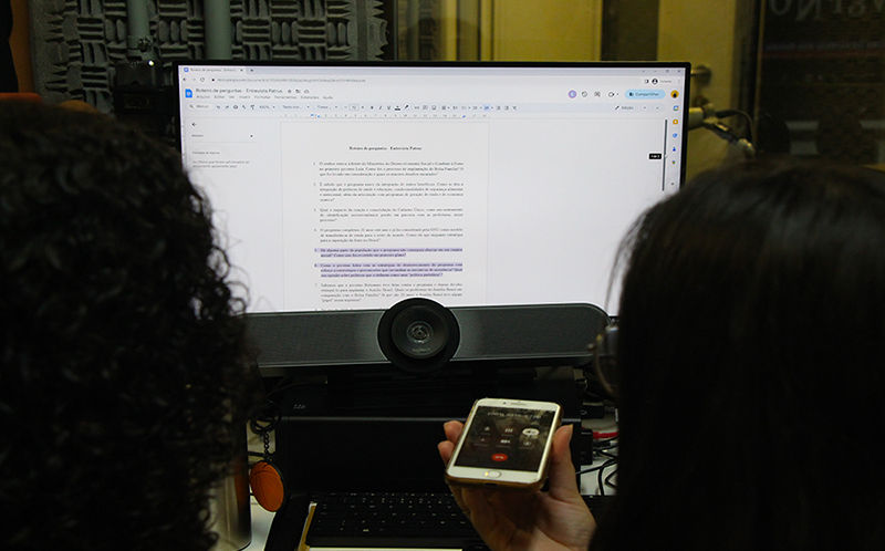 A foto mostra duas pessoas em frente a um computador com a tela aberta no documento com perguntas para a entrevista com o Deputado Patrus Ananias. Em ambos os lados, aparecem cabelos escuros, um cacheado e outro liso. Ao centro há um celular com a chamada em ligação.