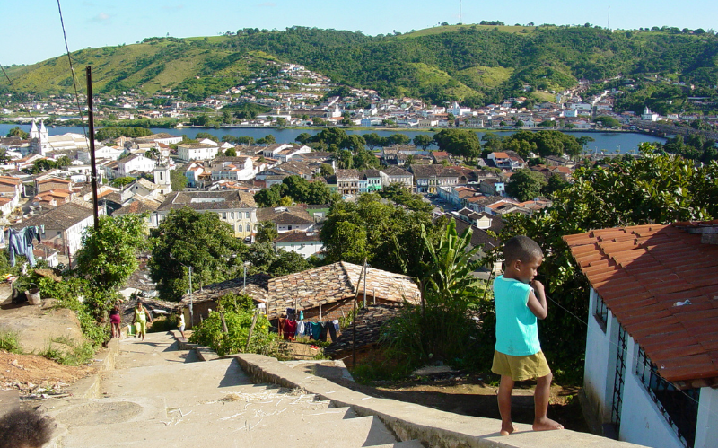 No primeiro plano, à direita, uma criança vestindo uma blusa azul; em segundo plano, casas de cores diferentes com diversas árvores espalhadas entre elas. À esquerda, há duas pessoas subindo uma longa escada por entre as casas. Mais ao fundo, há um rio que corta a cidade.