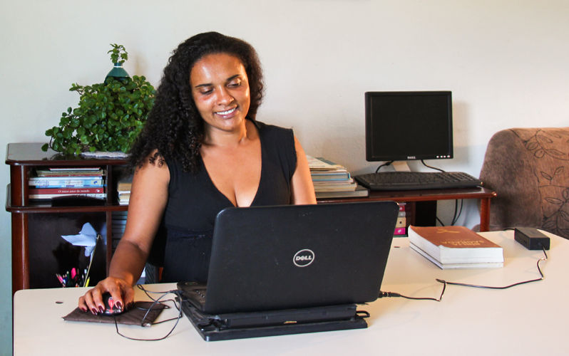 a imagem mostra uma mulher sorrindo. Ela tem cabelo castanho escuro, cacheado e está com um vestido preto. Ela está sentada em seu escritório, trabalhando em frente ao computador. Ao fundo, há um outro computador desligado em uma estante com livros e uma planta. No canto, aparece parte de um sofá.
