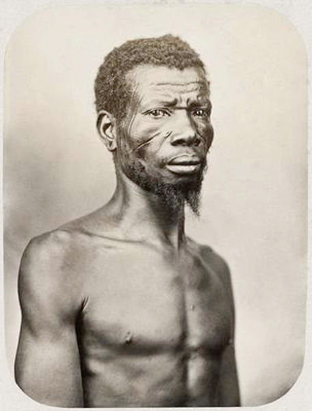 Um retrato fotográfico de busto, em preto e branco com tons de sépia, de um homem negro. Está posicionado em plano médio, com o olhar direcionado para o horizonte. Ele está com o torso nu, revelando uma constituição física magra e definida. Possui cabelos curtos e crespos e uma barba em estilo cavanhaque. O elemento mais marcante são as escarificações (marcas tribais) em seu rosto: duas linhas horizontais paralelas na bochecha direita e marcas semelhantes na têmpora.
