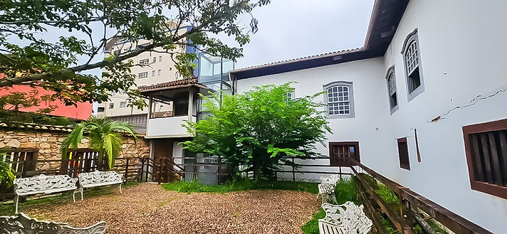 Pátio interno do Castelinho restaurado, com paredes brancas, janelas amplas de moldura cinza e telhado de madeira escura. No lado esquerdo da imagem, destaca-se um elevador externo de estrutura transparente, em vidro e metal, anexado discretamente à construção, permitindo acesso entre os pavimentos. 
