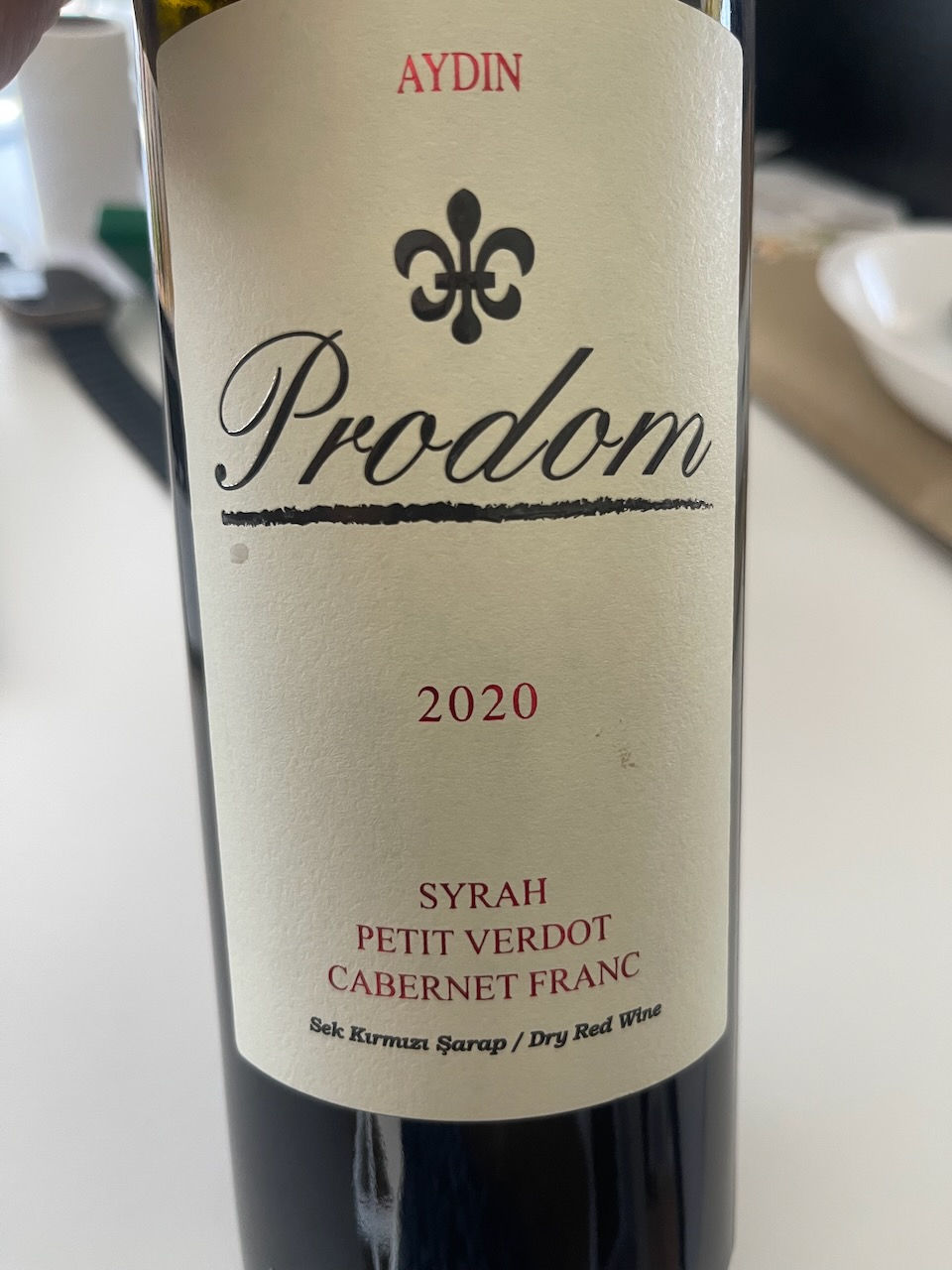 Wine bottle with label reading "Aydin Prodom 2020," featuring a fleur-de-lis. Varieties: Syrah, Petit Verdot, Cabernet Franc. White background.