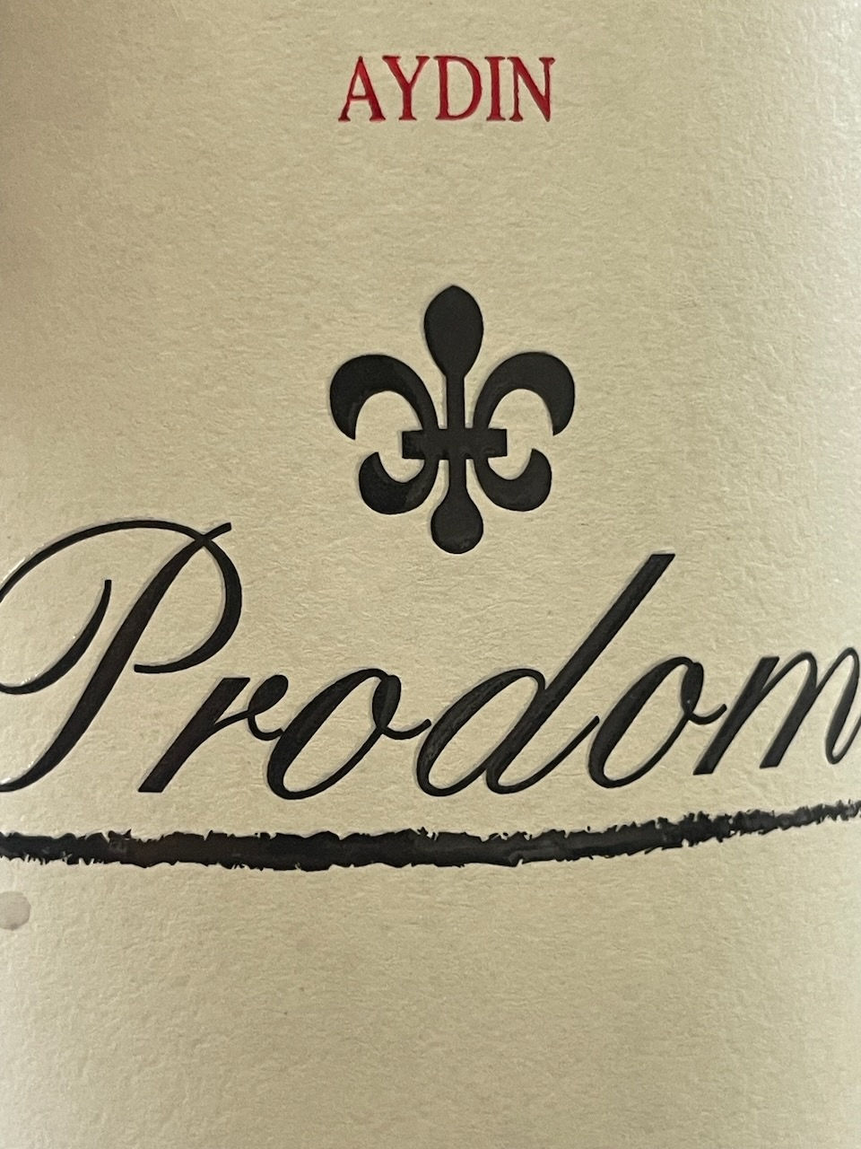 Close-up of a label with "AYDIN" in red, black fleur-de-lis, and "Prodome" in script. Cream background, elegant design.