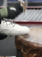 A gloved hand uses a hammer on a white shoe over a worn surface. Background shows a metal grid. Industrial setting.