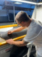 Man in white shirt cutting black fabric on yellow-edged table under bright light, focused work in an industrial setting.