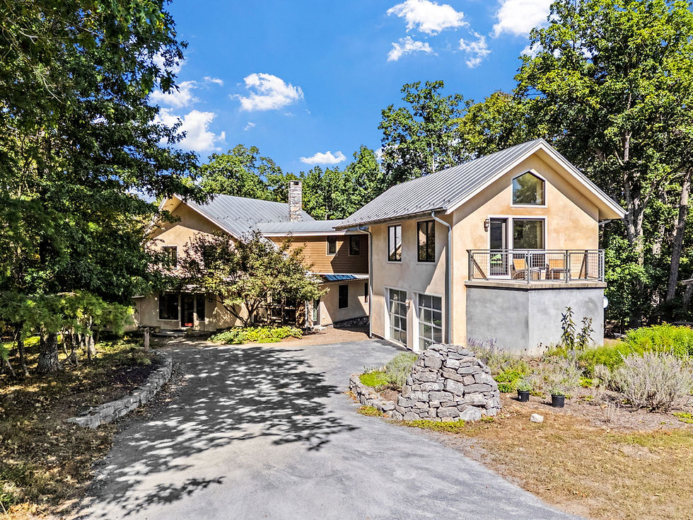 Modern beige house with a metal roof, surrounded by trees. A cobblestone path leads to the entrance. Sunny day with a blue sky.