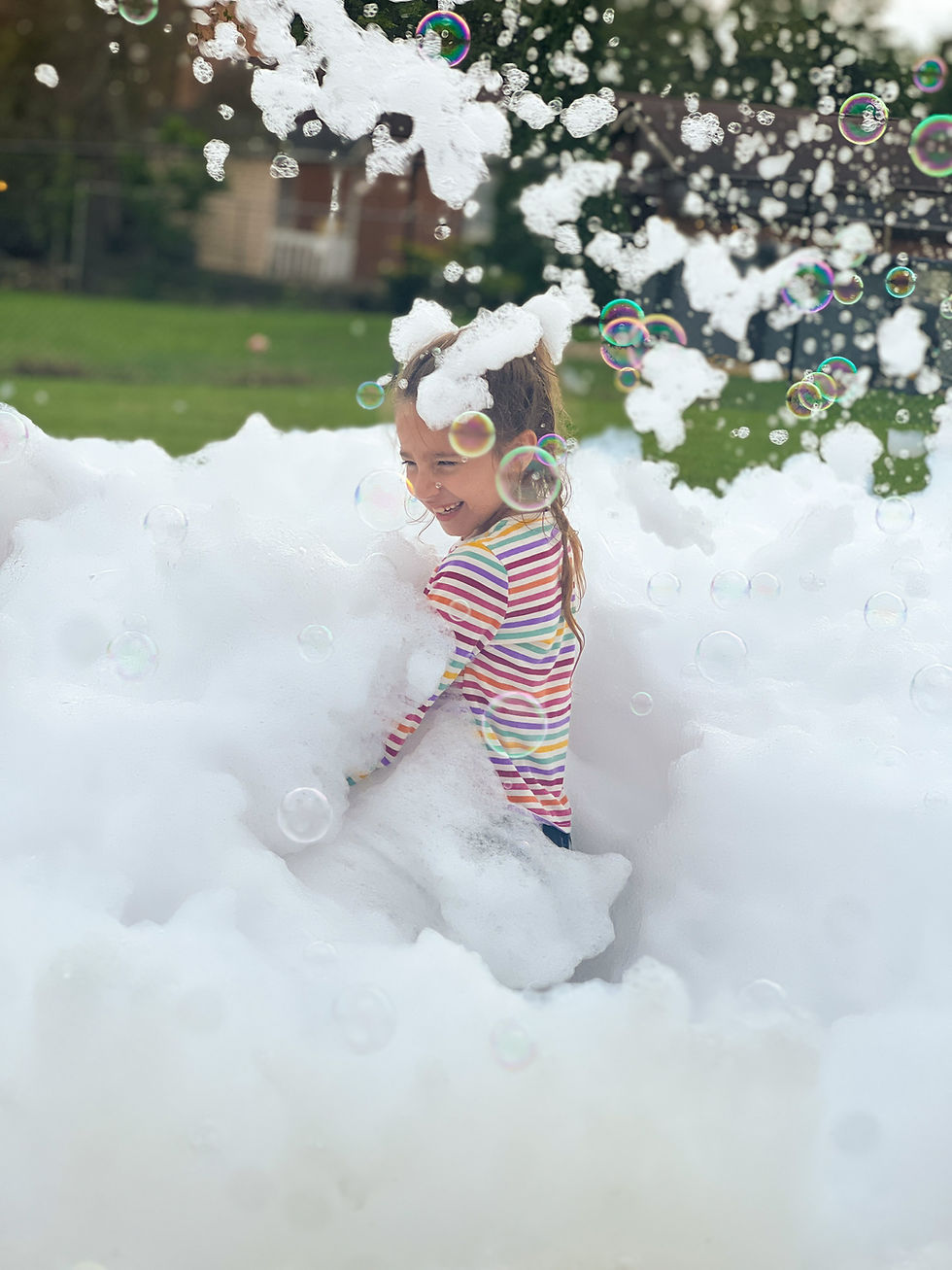 a young girl in a colorful striped shirt waist-deep in foam laughs as bubbles float overhead
