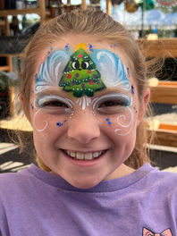 Child smiling with a festive Christmas tree face painting featuring snowy white details and holiday colors, professionally painted by Grand Creations by JE at a Tampa Bay holiday event.