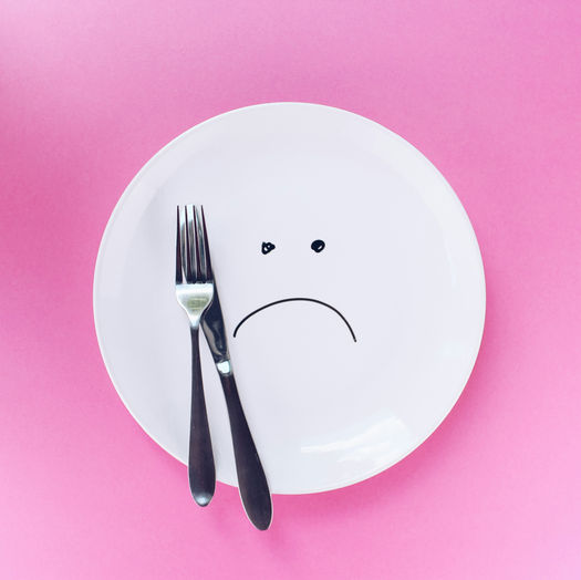 White plate on a pink background, with a simple sad face painted on the front with a black marker and a knife and fork on the right side