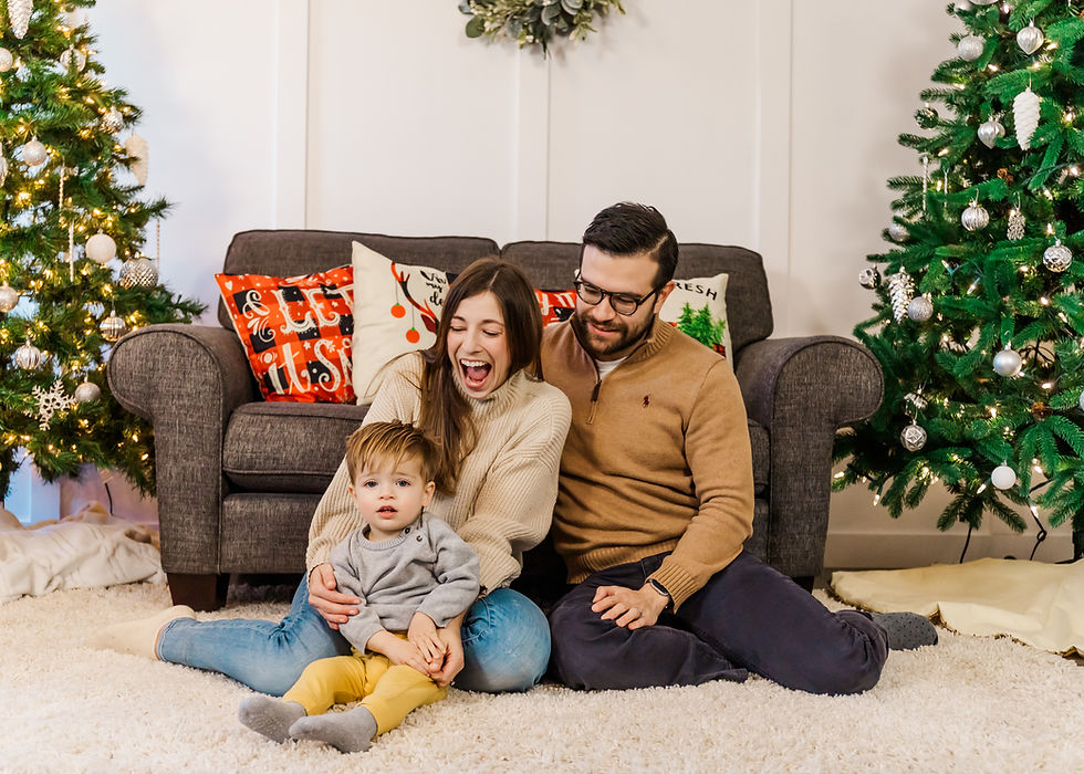 Family of three sitting on the ground during a Christmas Mini Session. 2 Christmas trees set up with a cozy couch and festive pillows. Mom laughing at toddler boy