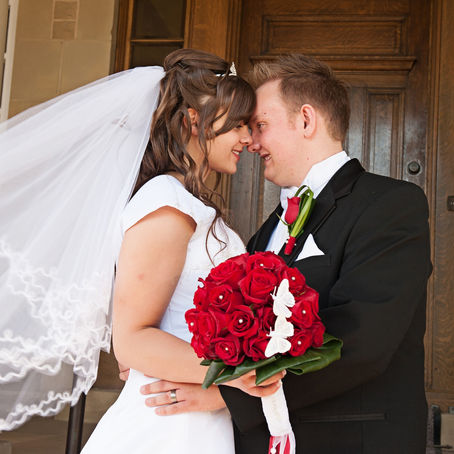 Wedding picture of a couple looking at each other and smiling. Veil flying behind the bride, large red rose bouquet with white butterflies in it visible