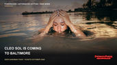 Cleo Sol rests both hands on her head while partially submerged in calm water near a shoreline.